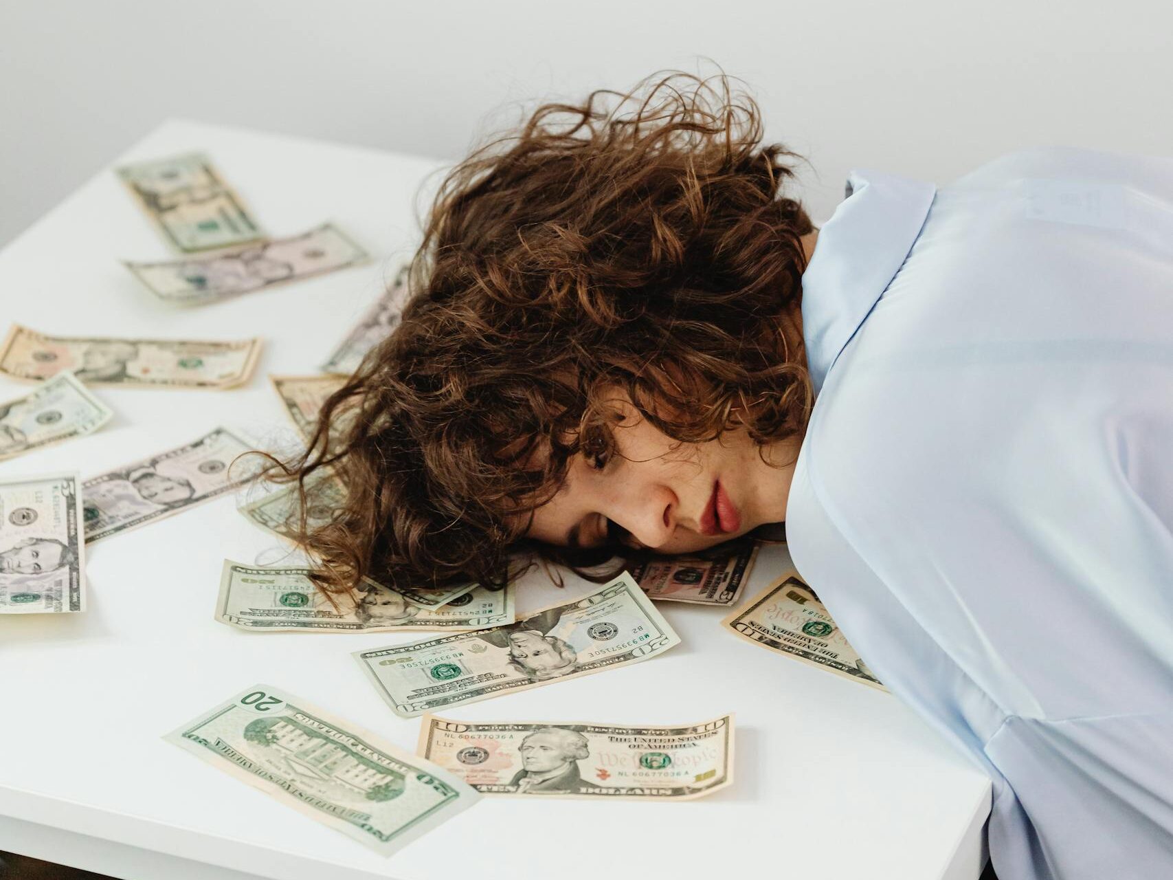 A woman with curly hair rests on a table covered in US dollars, expressing financial stress.